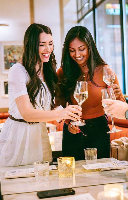 Two women standing at the head of a table at a restaurant doing a cheers with others at the table who's hands are on the shot